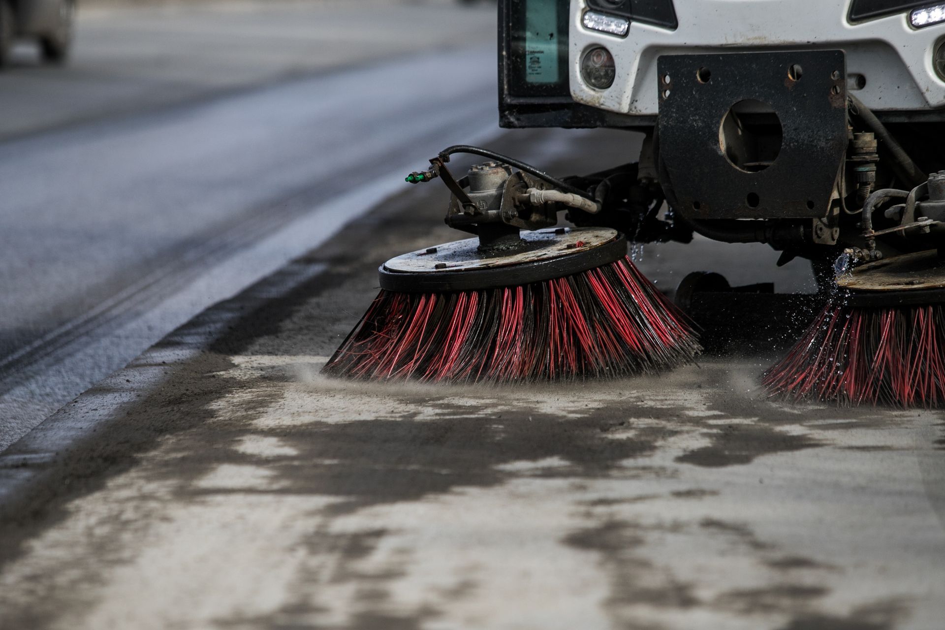 Street sweeper with brushes and water spraying on a road. A commercial sweeping vehicle cleaning a roadway curb with rotating brushes, removing dirt and debris from the pavement surface.