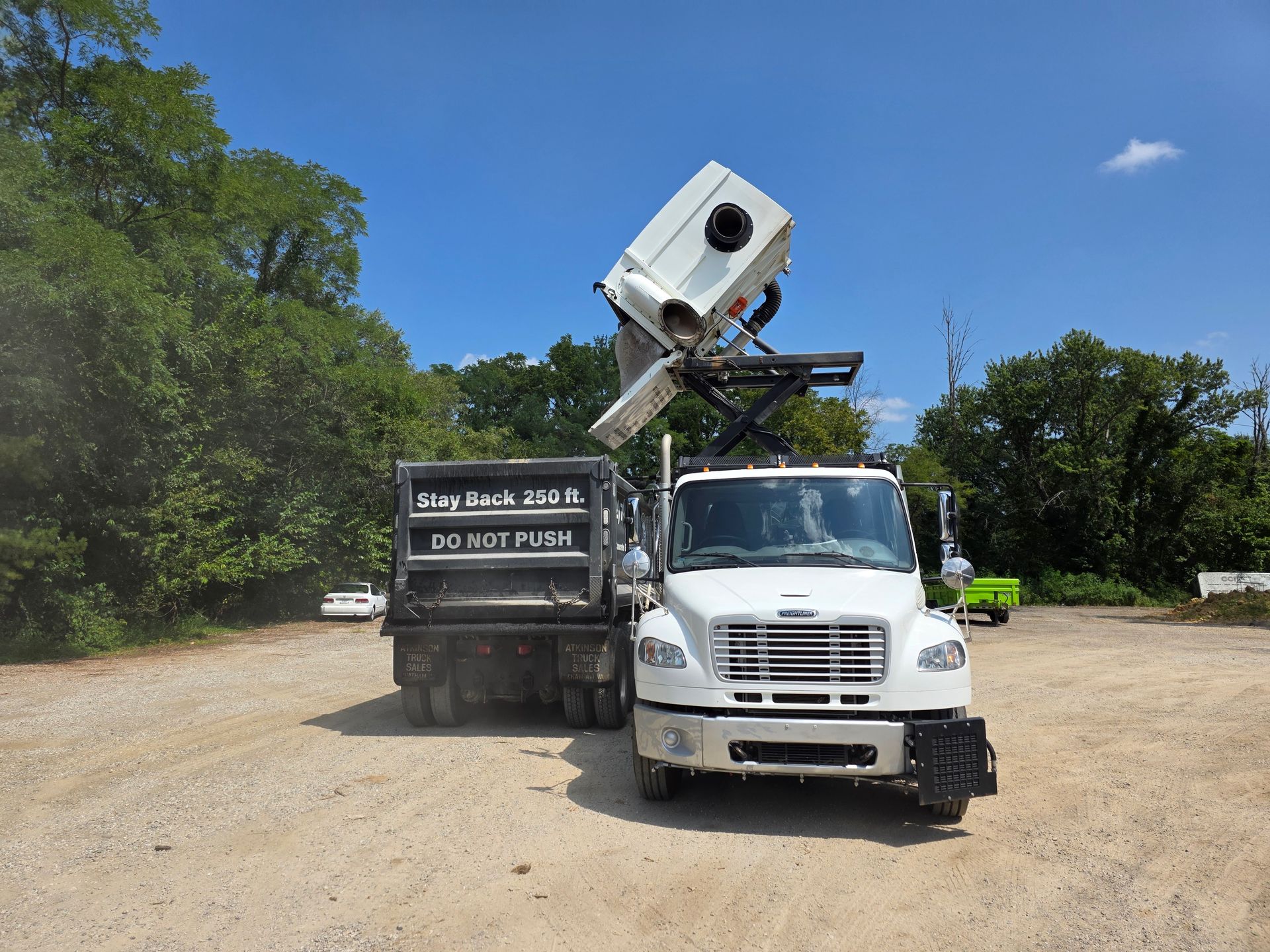 Street sweeper cleaning a brick-paved public park with benches and trees. White and dark gray vehicle.
