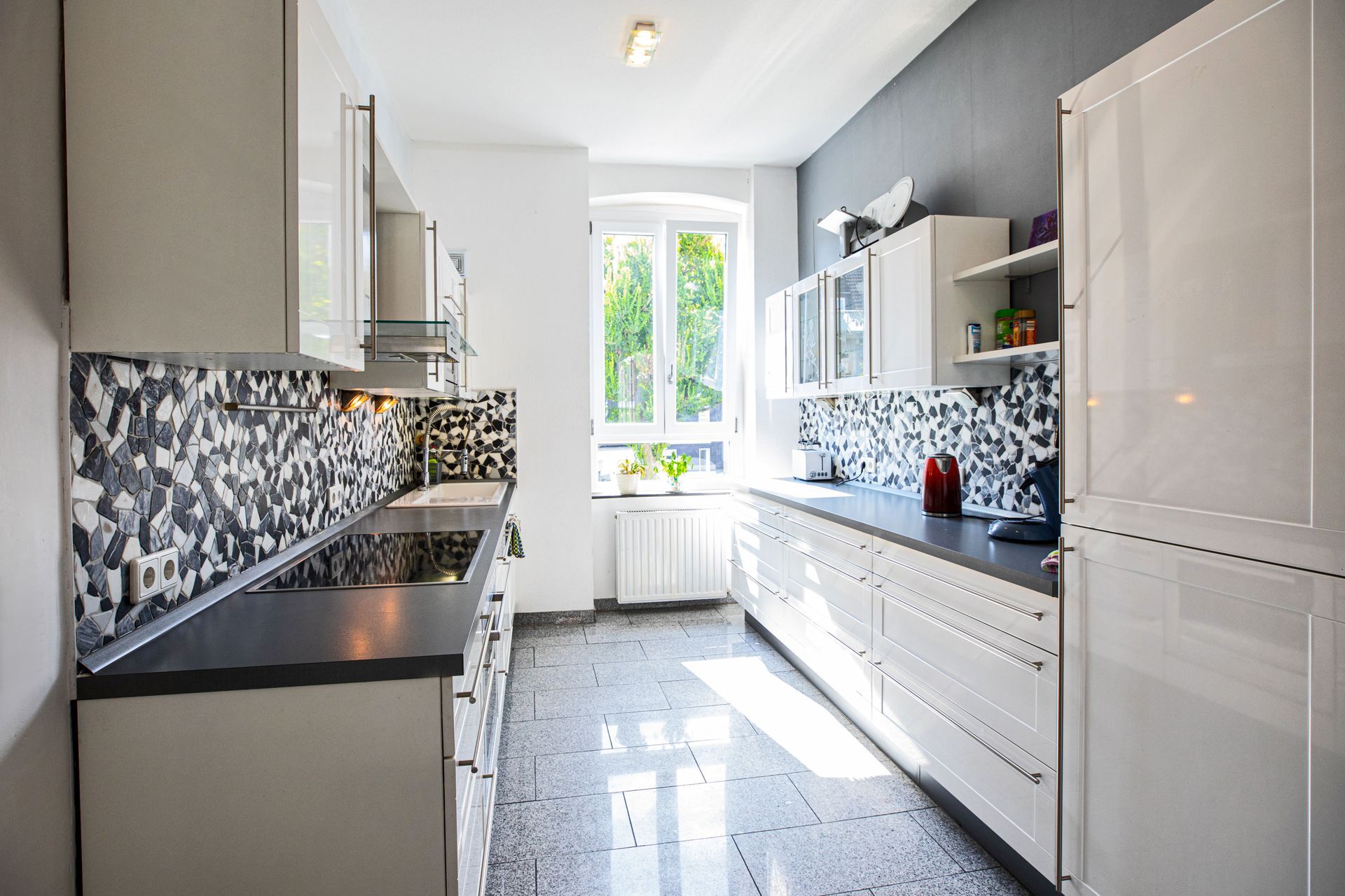Modern white kitchen with gray backsplash, stainless steel appliances, and window.