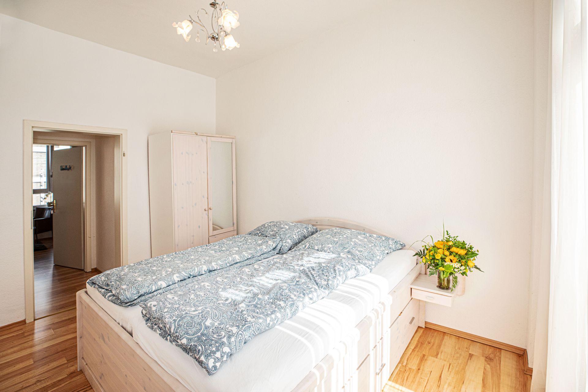 Bedroom with a bed, wardrobe, wooden floor, and a bouquet of yellow flowers on a side table.