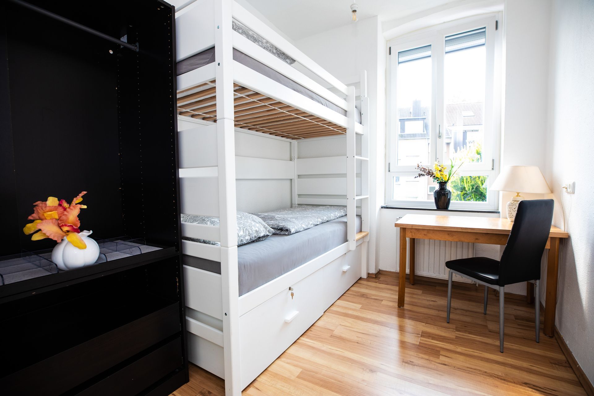 White bunk bed in a bright room with wooden floor, desk, chair, and a dark closet.