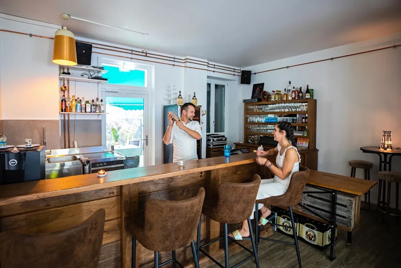 Bartender mixing a drink at a wooden bar for a seated woman. Interior shot with bottles and shelves visible.