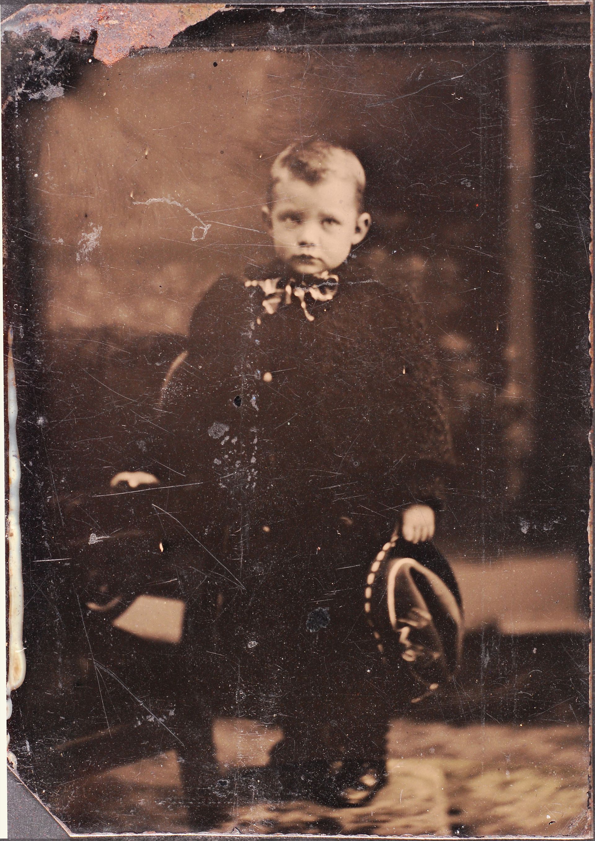 An old, sepia coloured photo of a young boy in a suit and holding his hat.