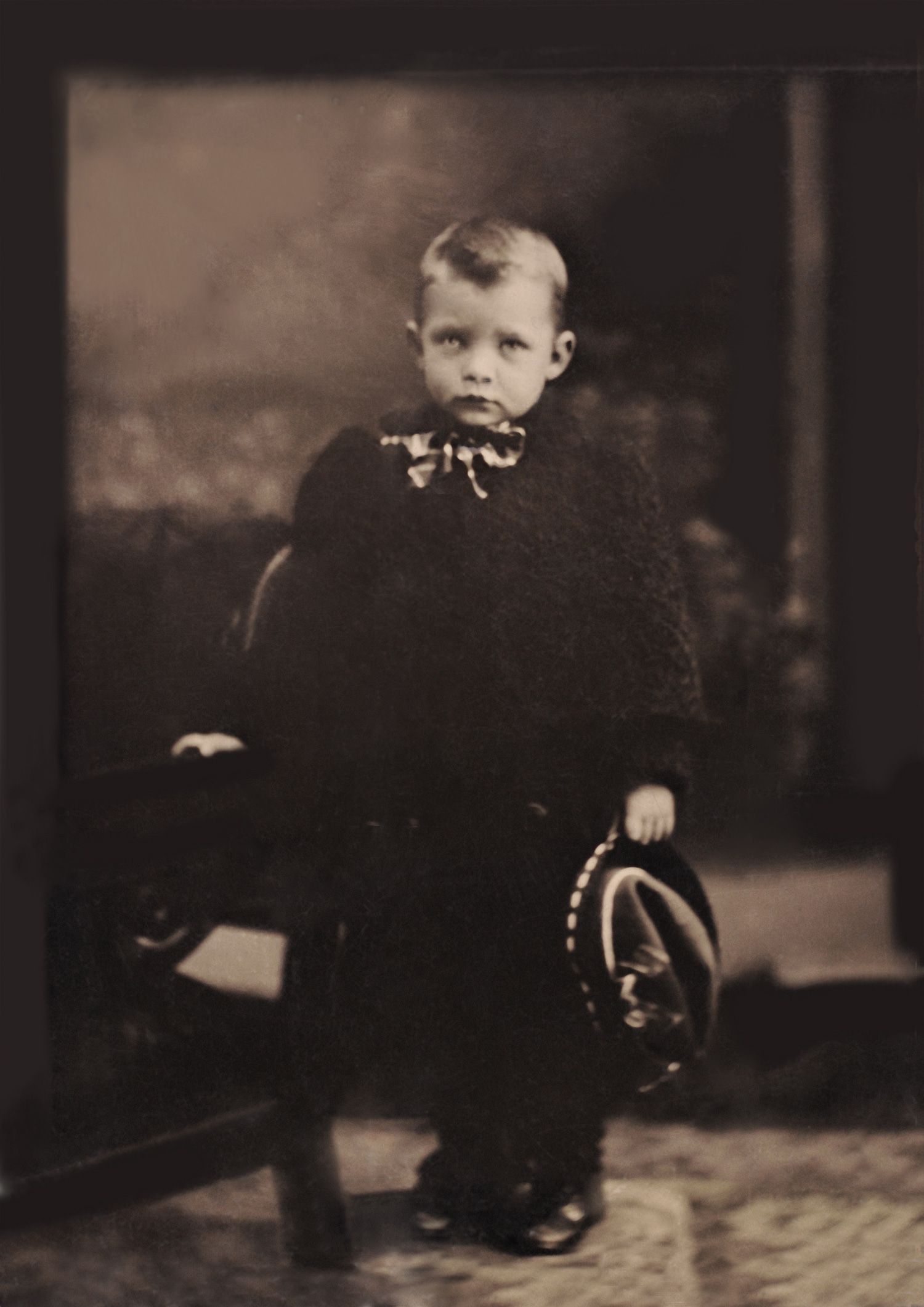 An old, sepia coloured photo of a young boy in a suit and holding his hat.