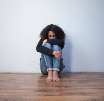 A person sitting on a wooden floor, hugging their knees with their head down against a plain white wall.