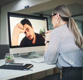 A person with gray hair in a ponytail sits at a desk viewing a video call with a man who has his hand on his forehead.