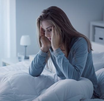 A person sitting on a bed in a dimly lit room, holding their head with both hands with a distressed expression.