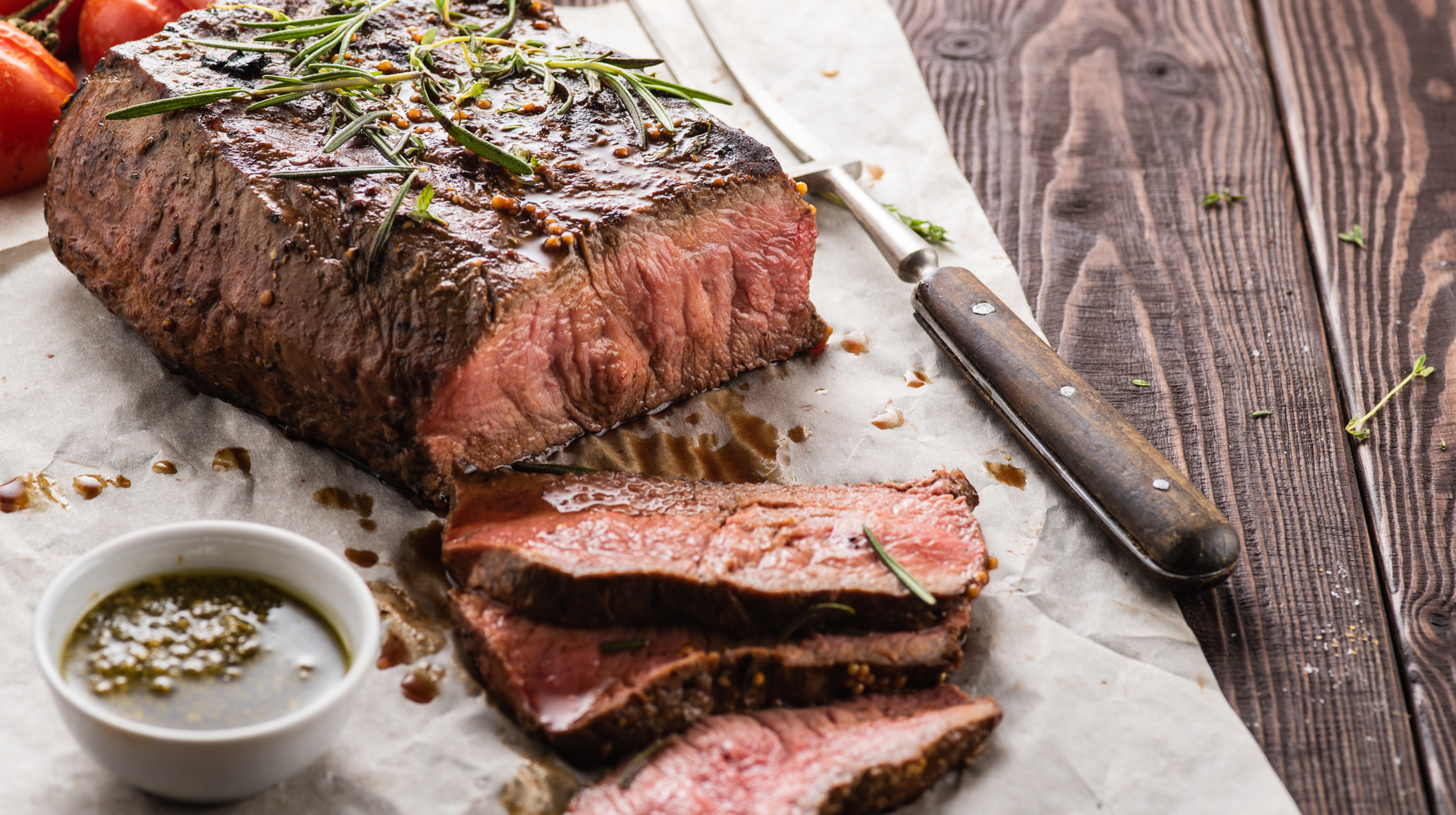 A large piece of steak is sitting on a wooden table next to a bowl of sauce.