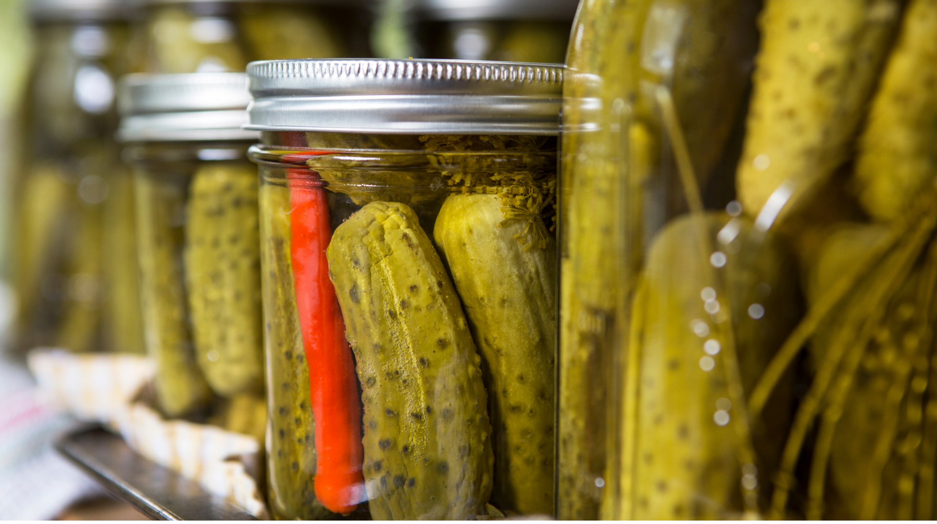 A row of jars filled with pickles and a red pepper.