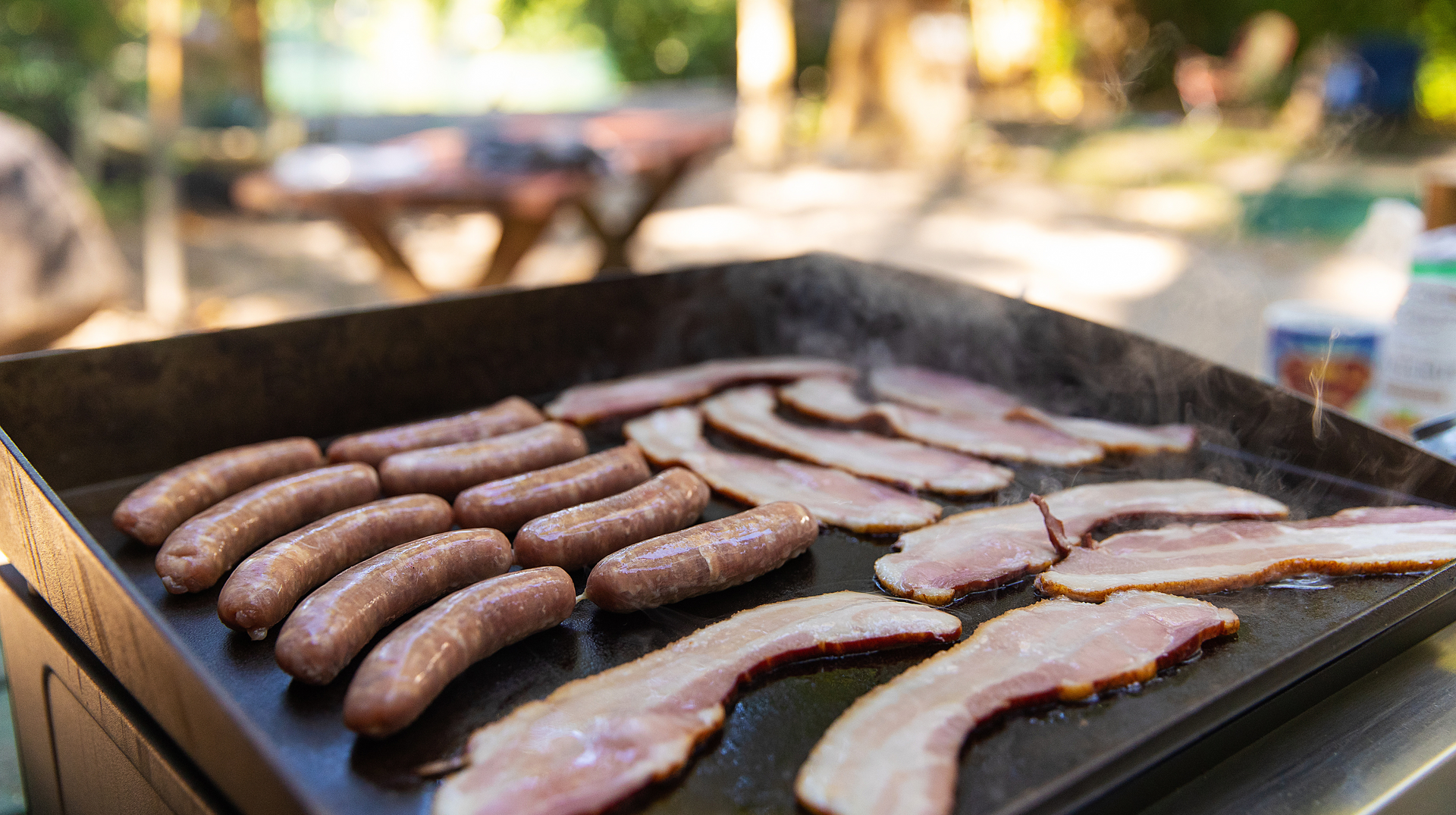 Sausages and bacon are cooking on a grill outside.