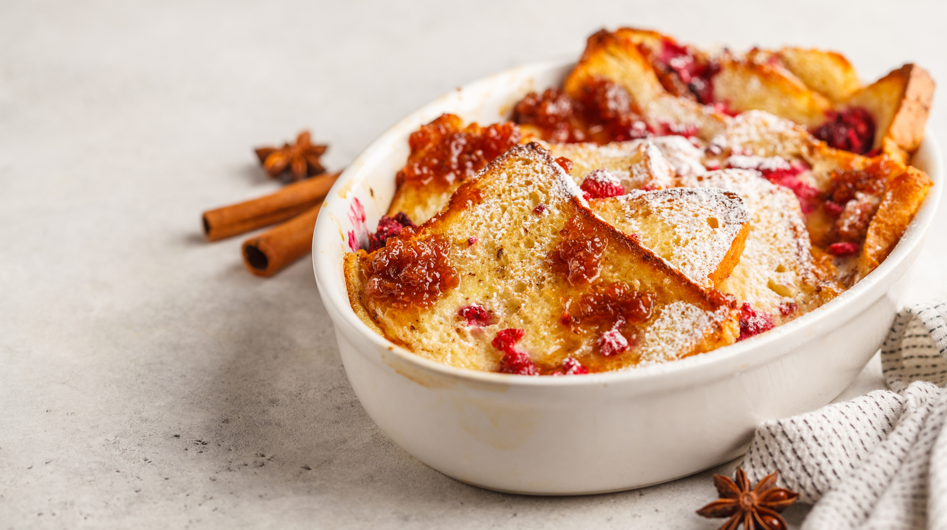 A bowl of bread pudding with cranberries and cinnamon sticks on a table.