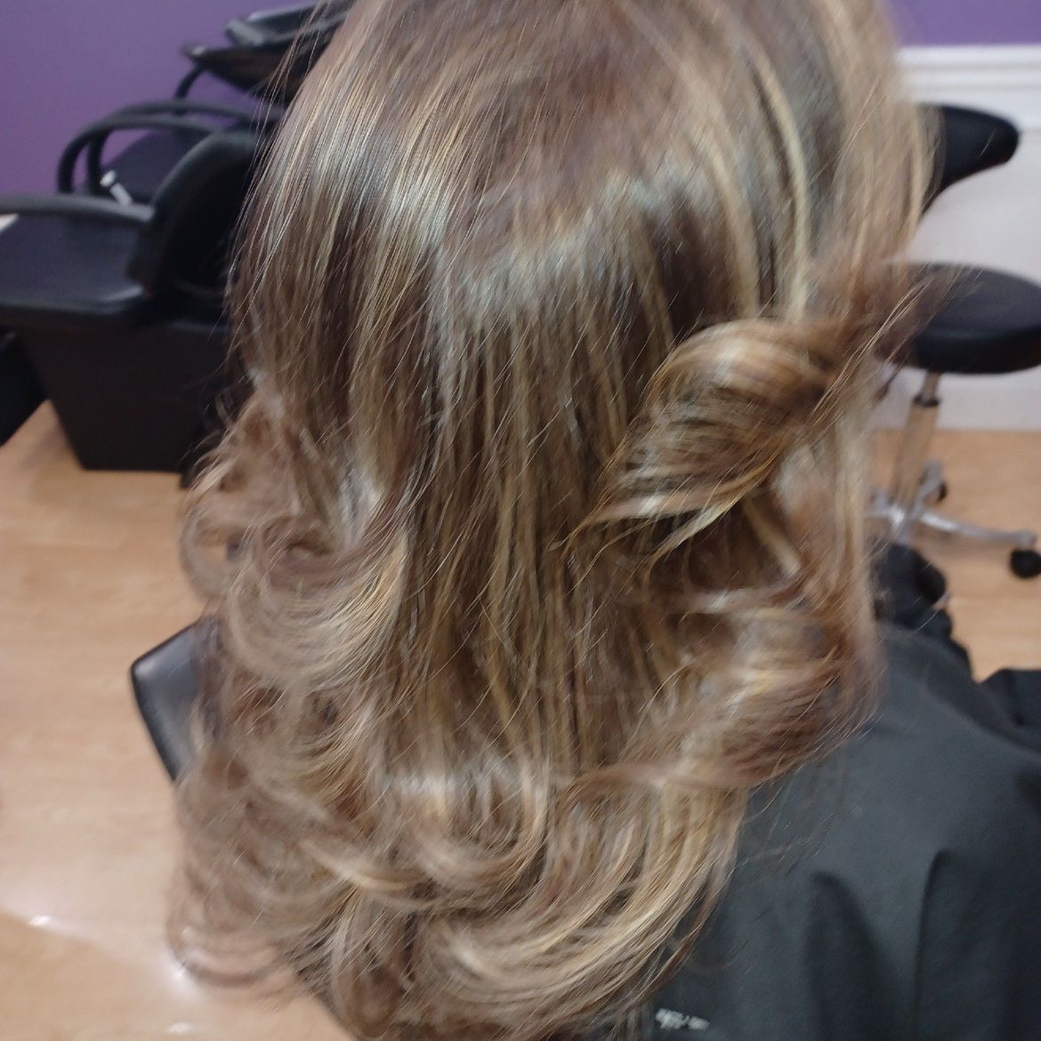 A woman with long hair is sitting in a chair in a salon with a curled style.