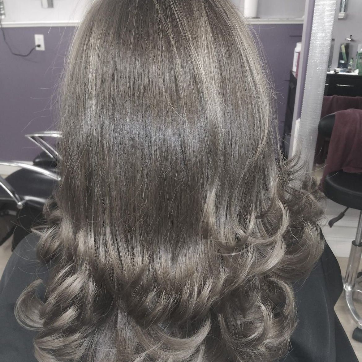 A woman with long gray hair is sitting in a chair in a salon, after having a curled style.