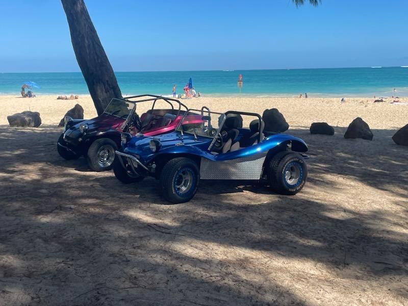 Two buggies are parked on the beach under a tree.