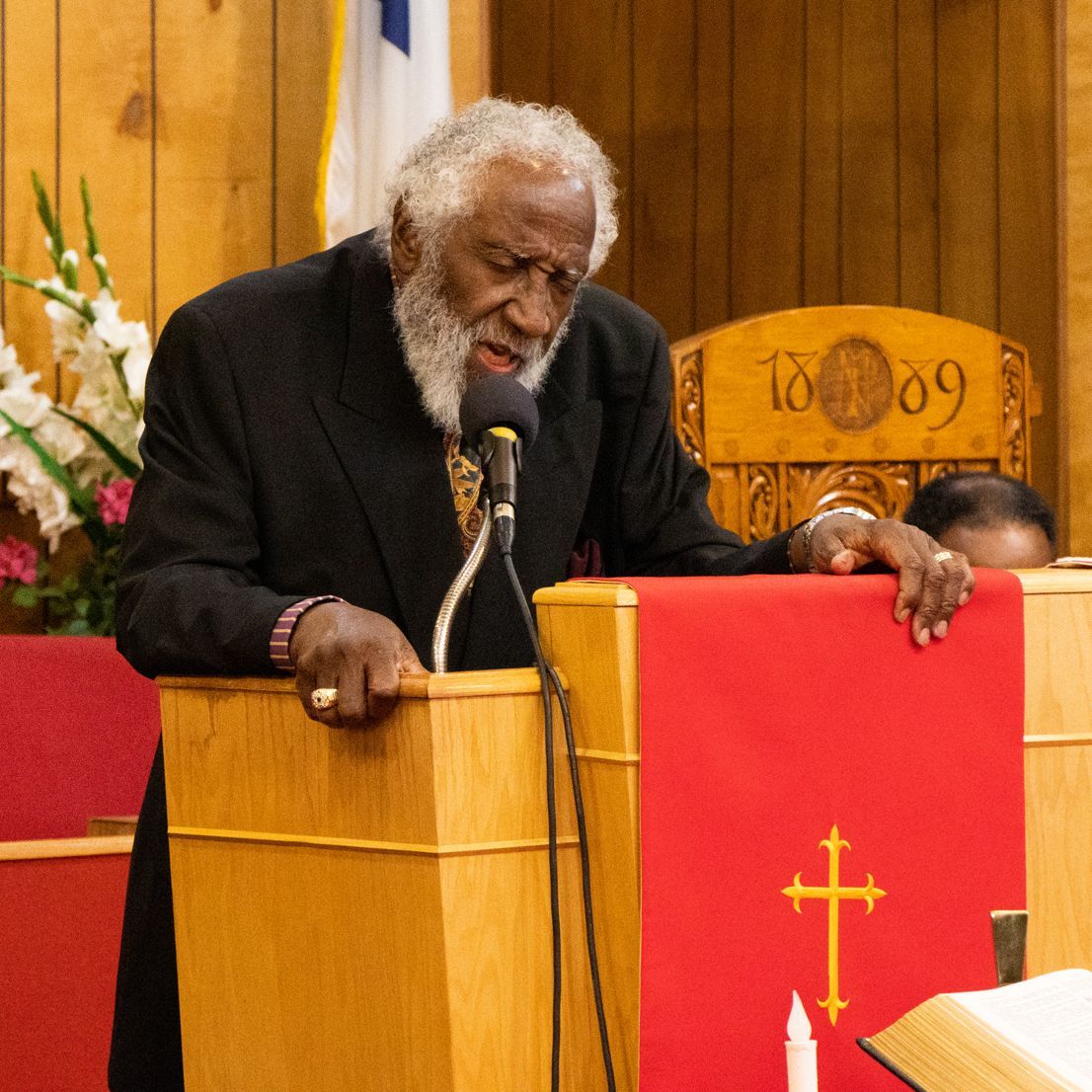 Pastor Ballenger at the pulpit praying, with 1889 chairs in the background.