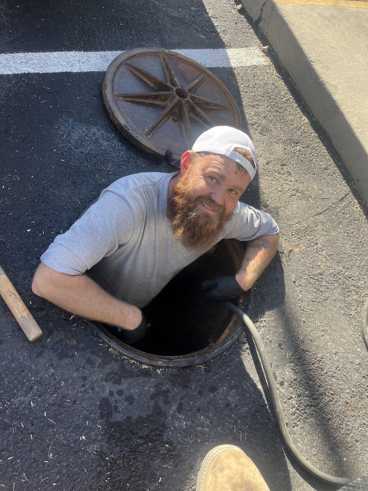 Man in a manhole, smiling, wearing a baseball cap and light gray shirt, working with a hose.