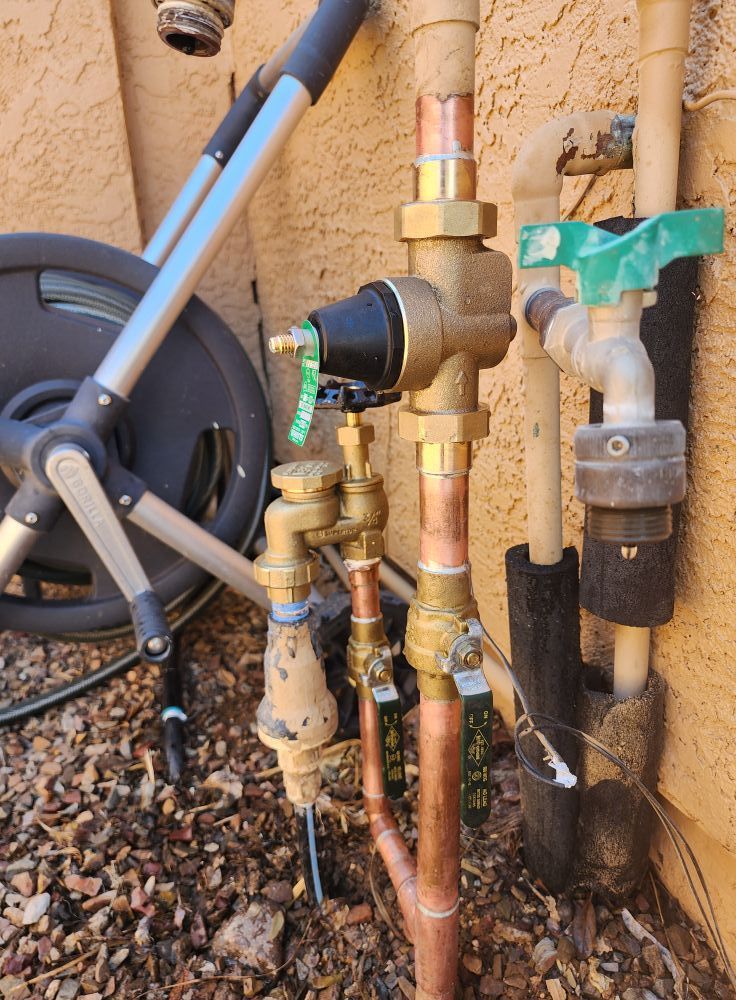 Outdoor water pipes with various valves and fittings attached to a stucco wall, with a hose reel in the background.