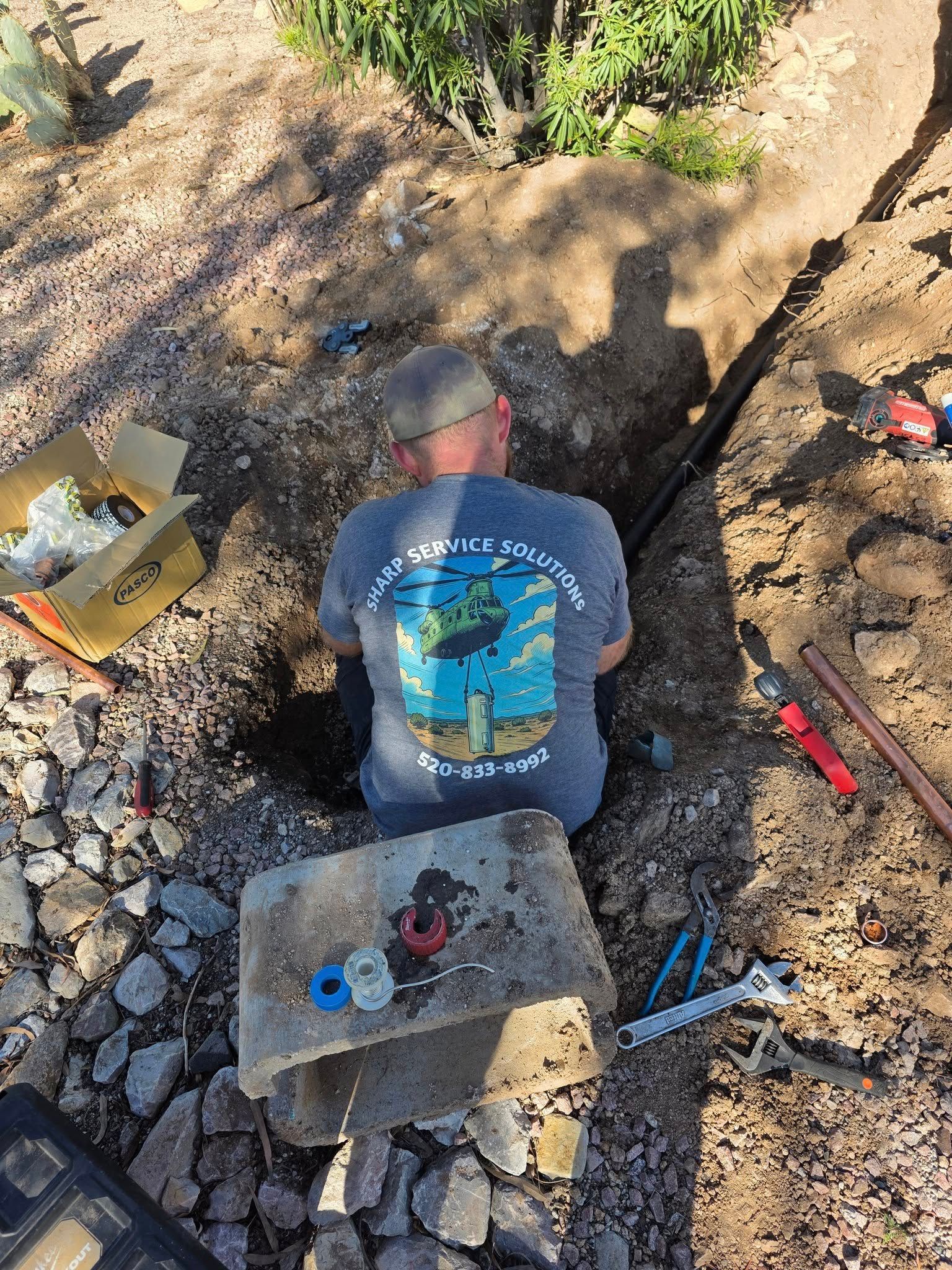 Man working in a ditch, with tools and pipe, in an outdoor setting.