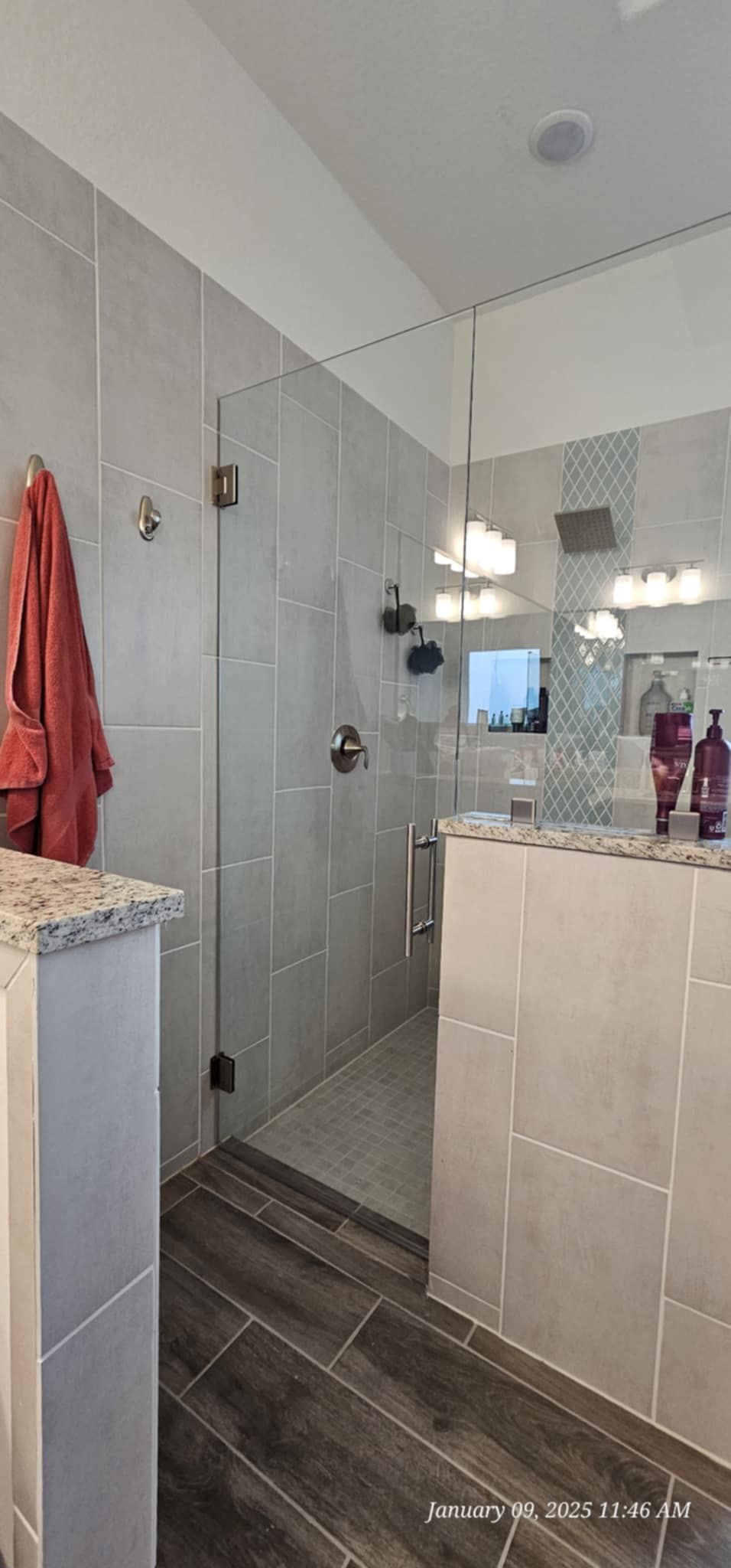 Modern bathroom with glass shower, gray tiles, and dark wood-look floor. A red towel hangs on a wall.