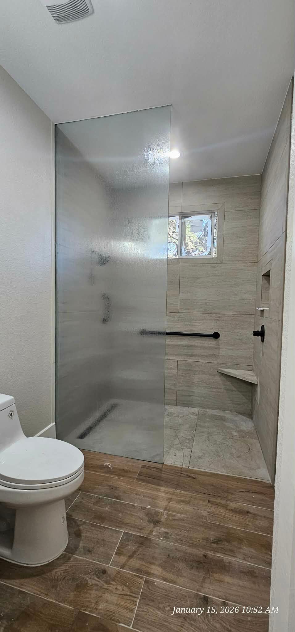 Bathroom with shower, toilet, and wood-look tile floor. A clear glass shower has a stone tile back wall with a window.