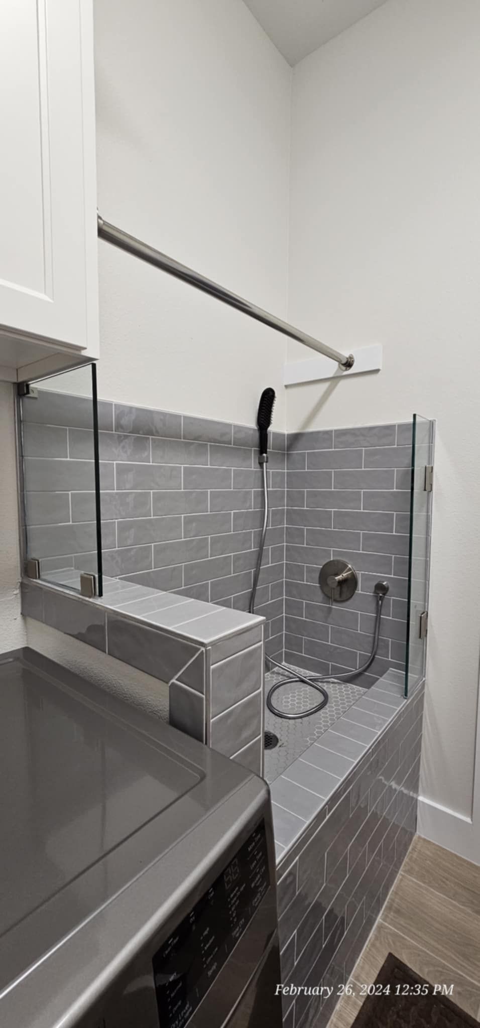 A modern tiled shower with a glass enclosure. A washing machine is visible in the foreground.