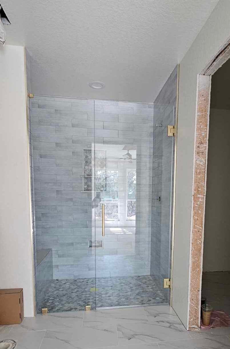 Frameless glass shower enclosure with brass hinges and handle, gray tile wall, and pebble floor.