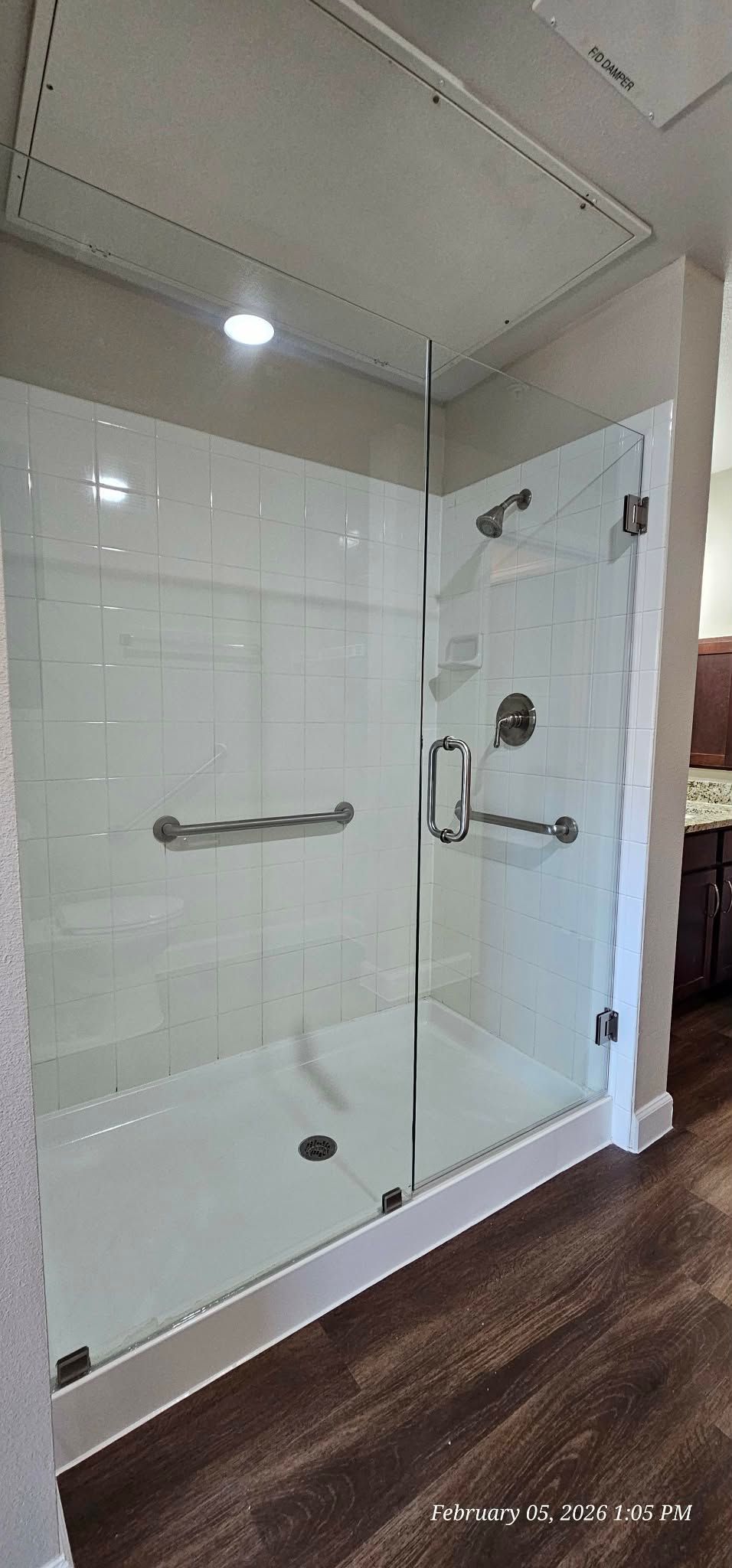 Glass shower with white tile walls and grab bars, on dark wood flooring.