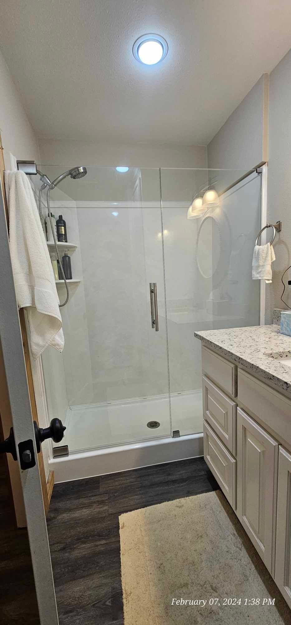 Bathroom with a glass shower, white vanity, and dark wood-look floor. A white towel hangs on the door.