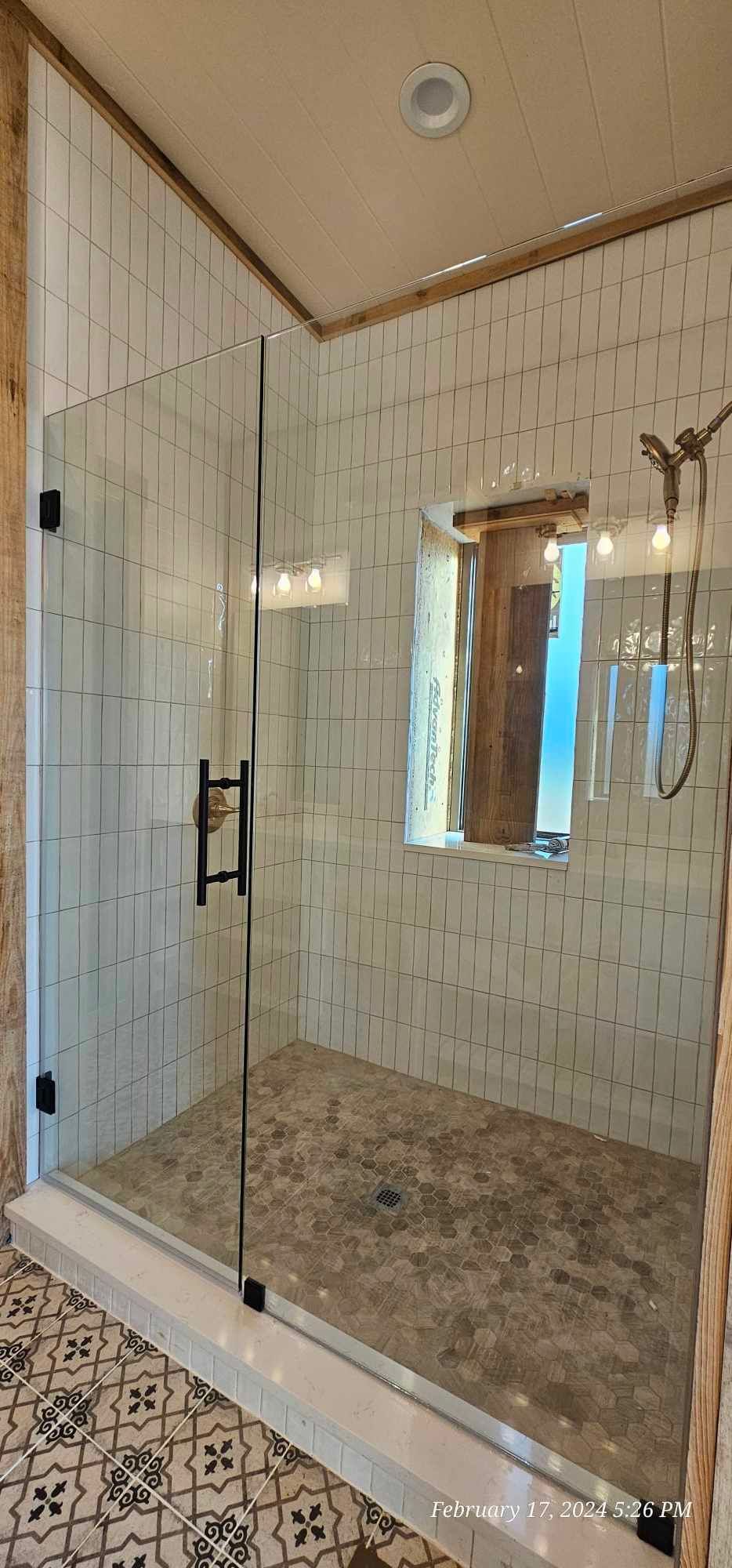 A glass shower enclosure with black hardware, white tile walls, and a patterned tile floor.