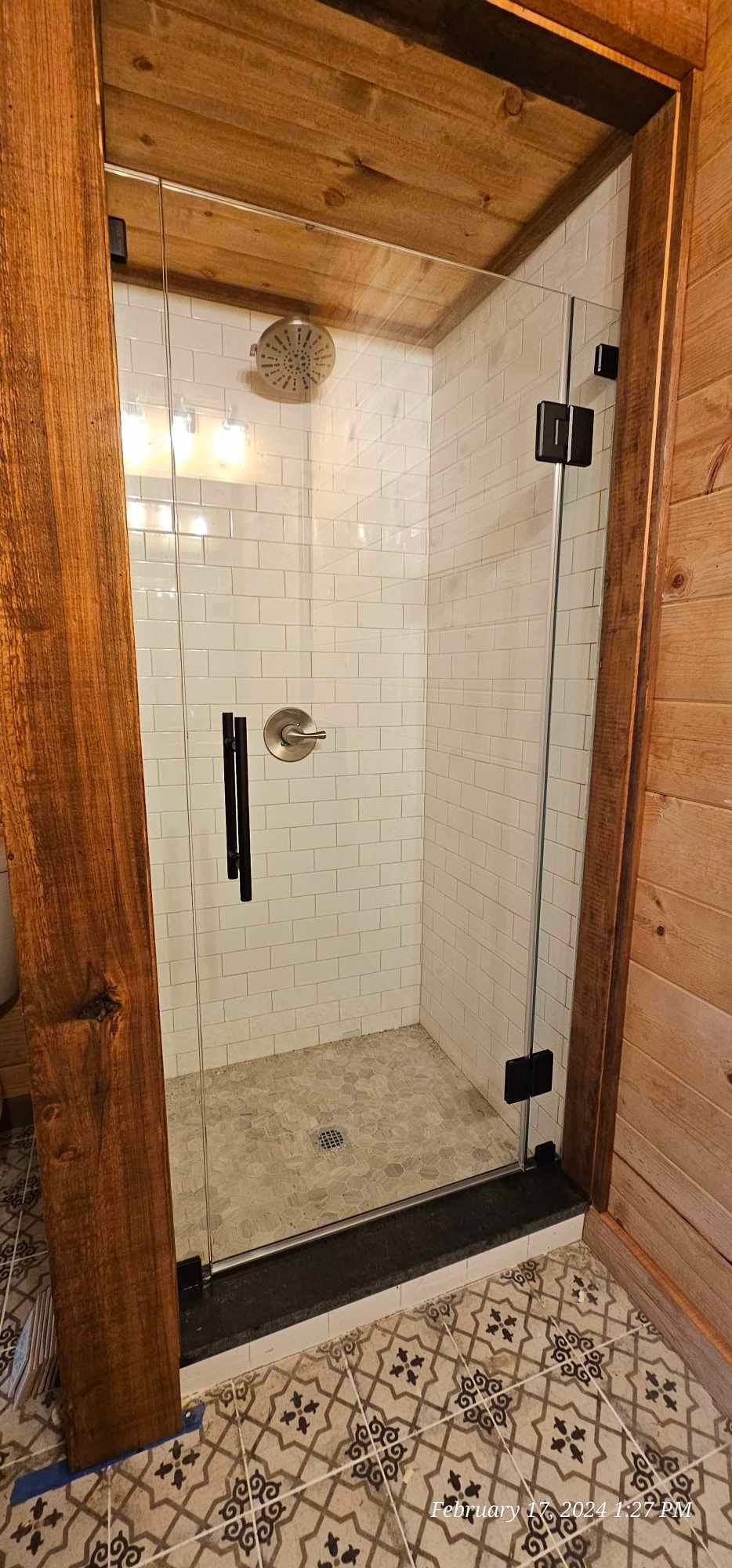 Shower stall with glass door and black fixtures, inside a rustic, wood-paneled bathroom.