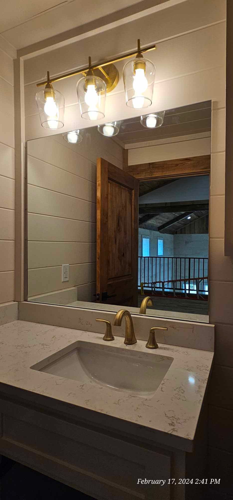 Bathroom vanity with a gold faucet and light fixture. A wooden door is reflected in the mirror.