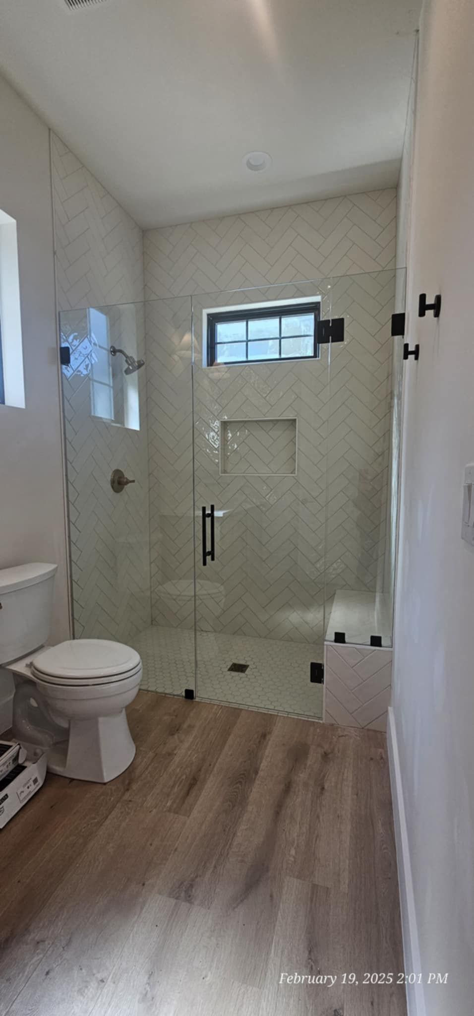 Bathroom with glass shower, toilet, and wooden floor. White tile shower walls with a small window.