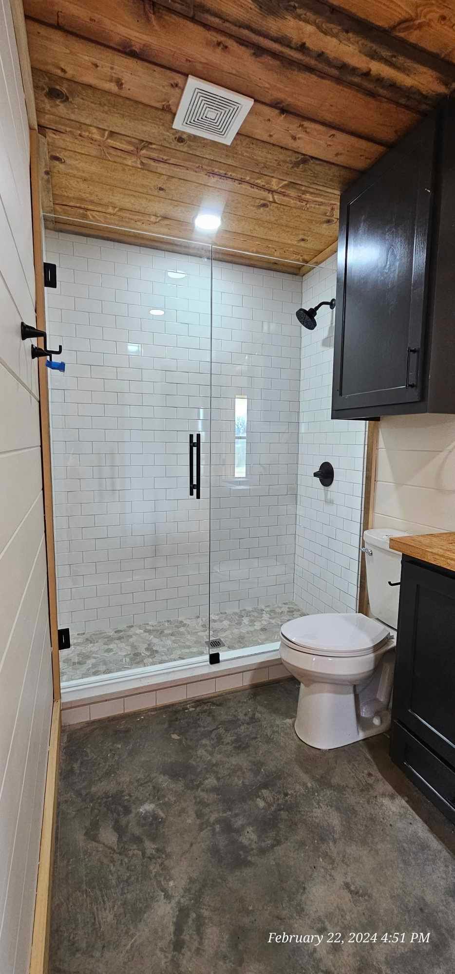 Bathroom with glass shower, white tile, black fixtures, wooden ceiling, and concrete floor.