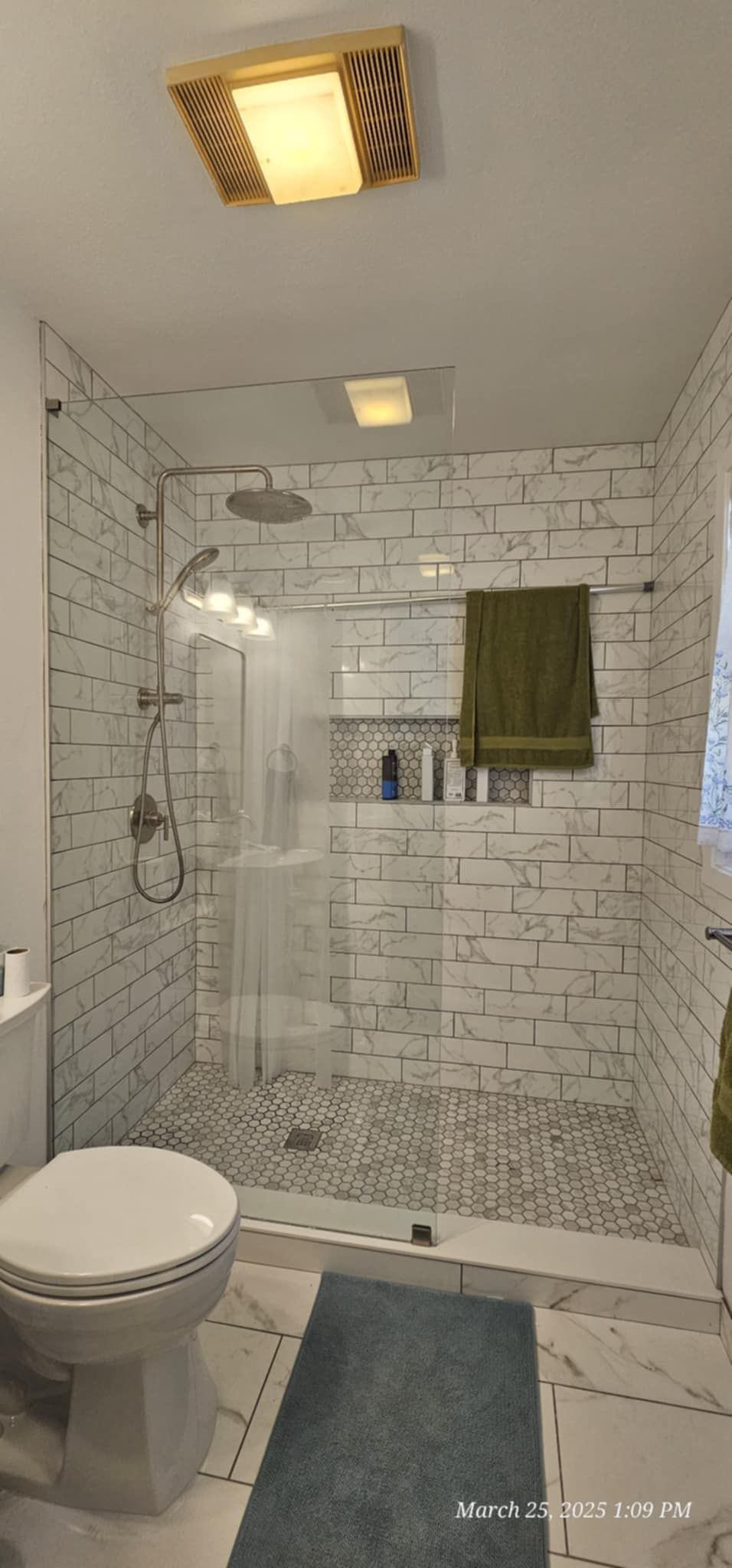 A bathroom with a toilet, shower, and green towel. White tile walls and a blue rug.