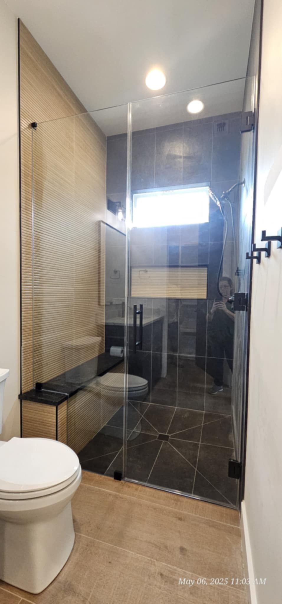 A modern bathroom with a glass shower, toilet, and patterned wall tile. The floor is light wood.