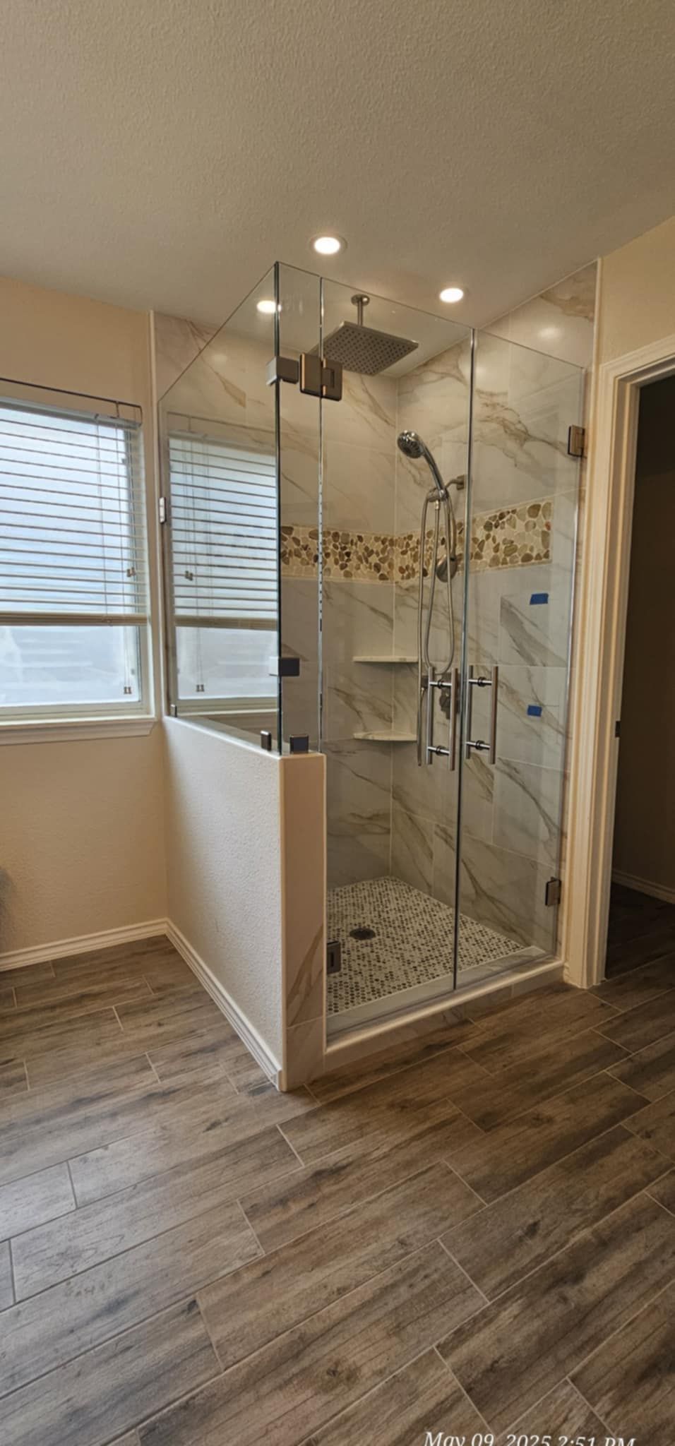 Bathroom with glass shower, marble tile, and wood-look flooring. Window with blinds on the left.