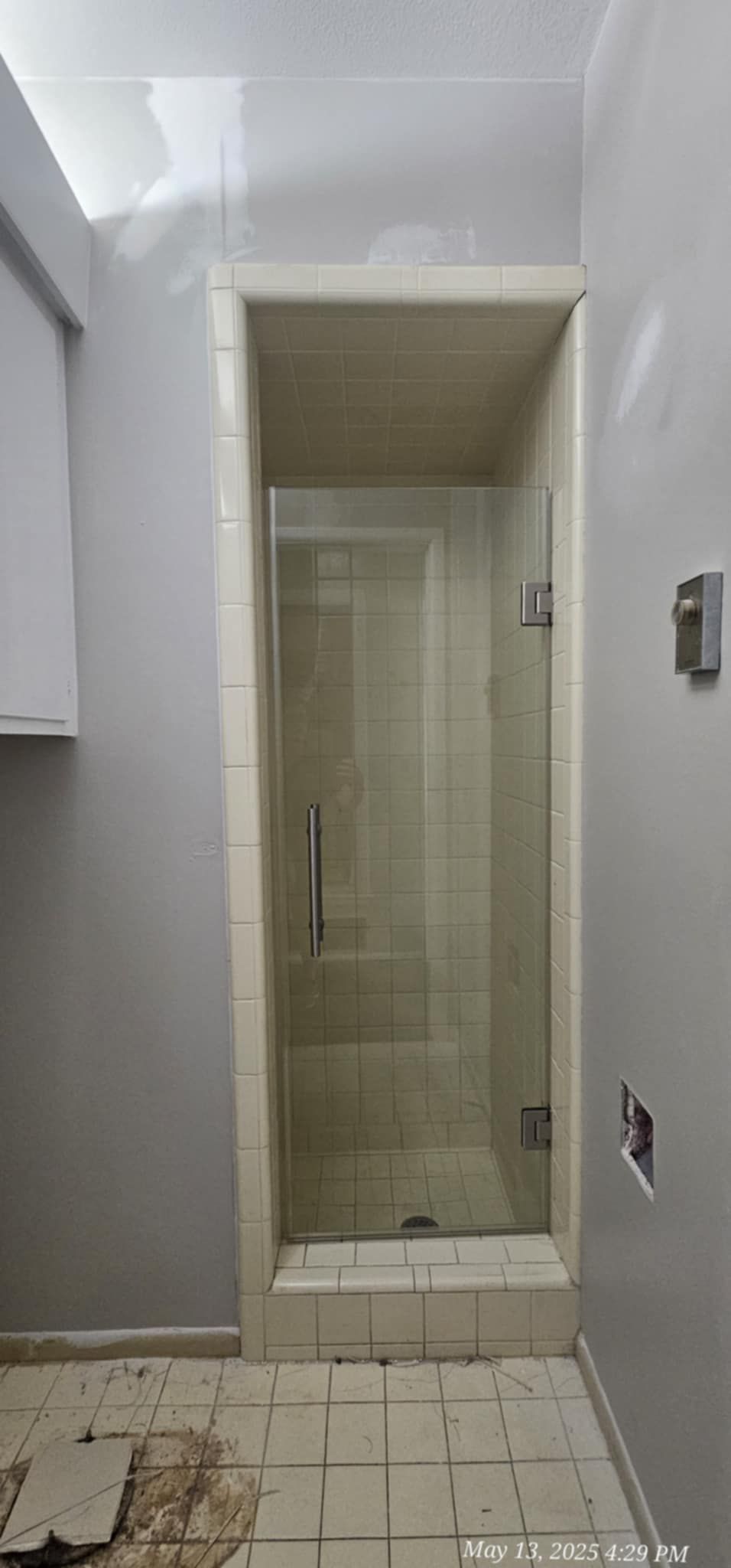 A shower with a glass door, framed in light-colored tile, stands in a room with gray walls and a damaged floor.