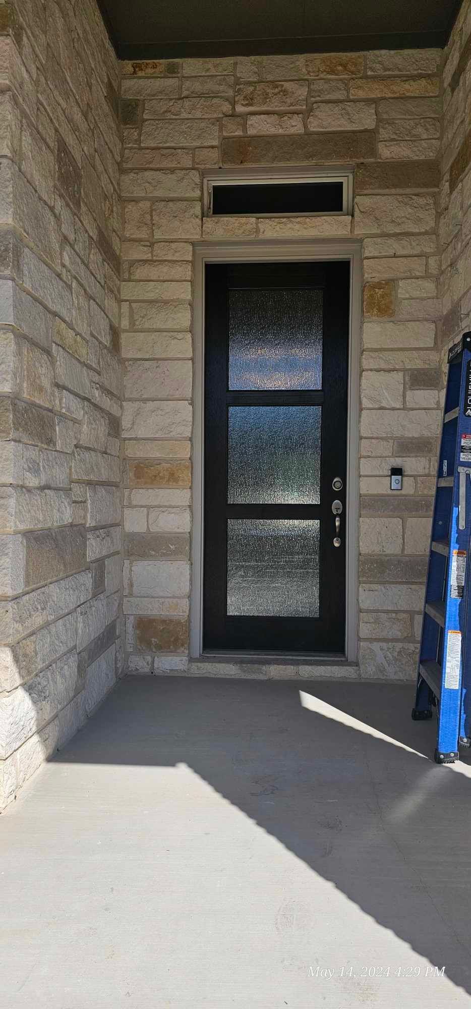 Black front door with three glass panels, stone exterior. Blue ladder visible on the right.