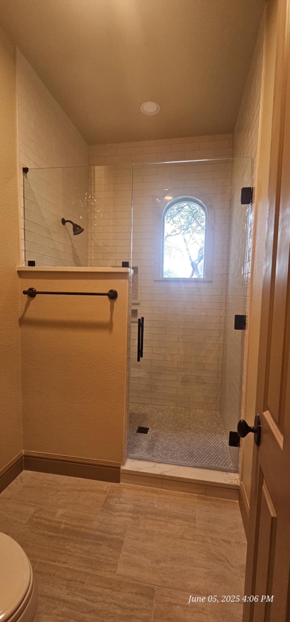 A small bathroom with a glass shower and an arched window. The walls are a light beige, the floor a tan color.