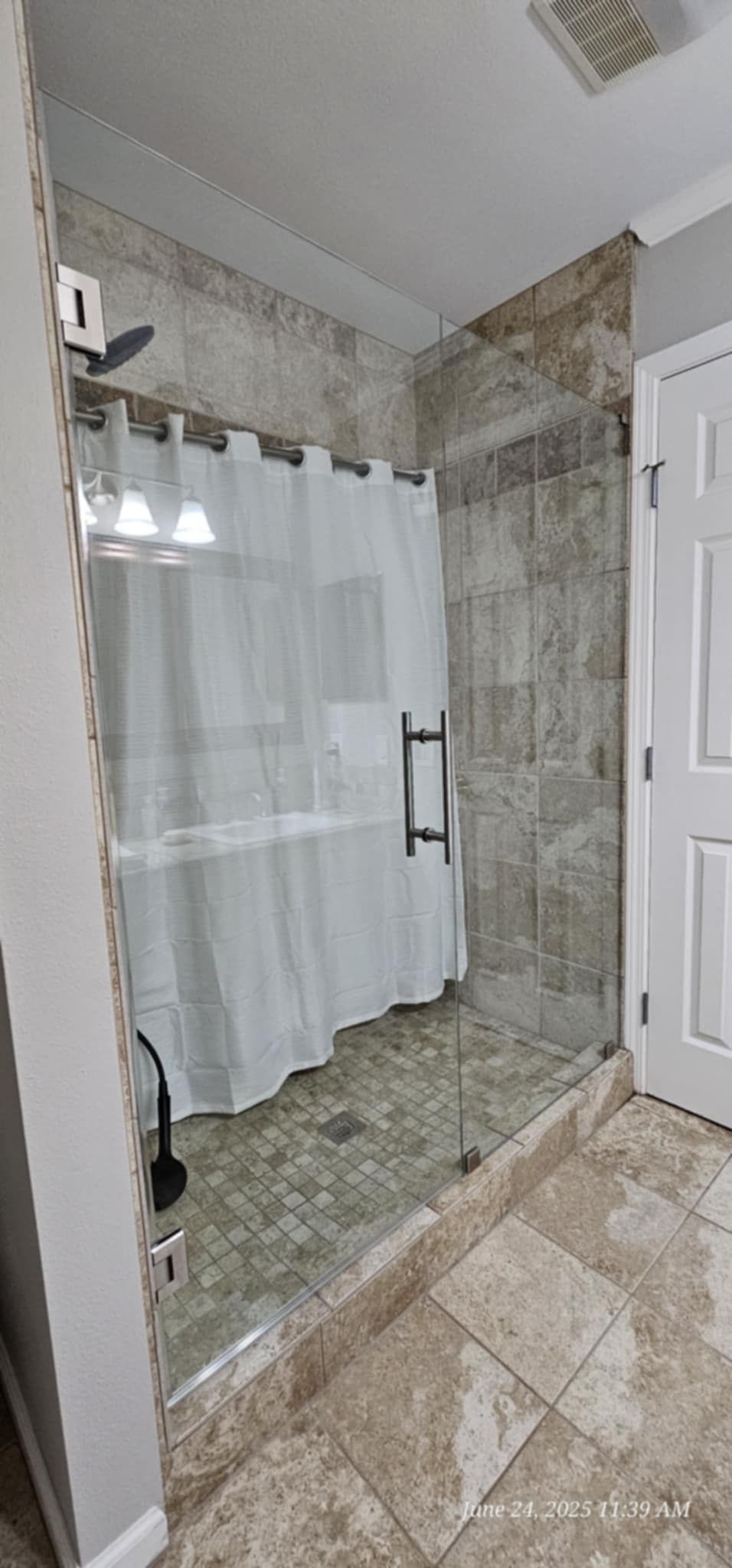 Bathroom with glass shower door, stone tile walls and floor, white shower curtain.