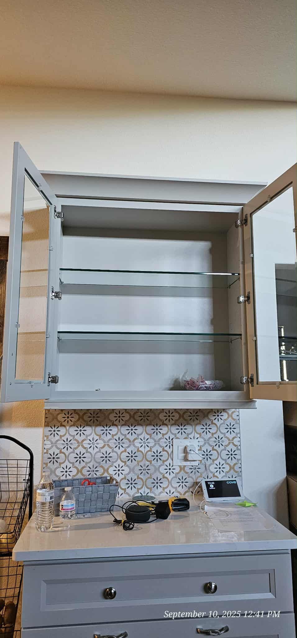 An open medicine cabinet above a gray cabinet. Glass shelves inside. The cabinet is above a tiled backsplash.