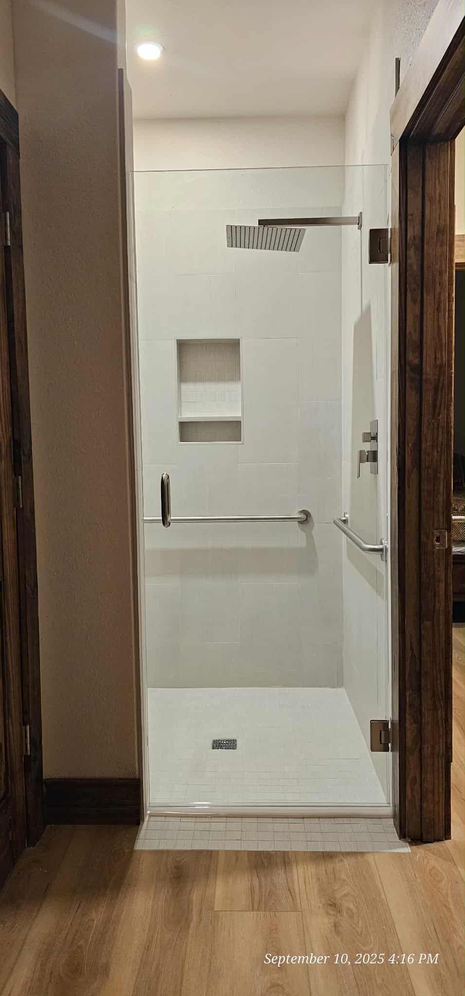 A walk-in shower with a clear glass door. The interior is white with a built-in shelf and a silver handrail.