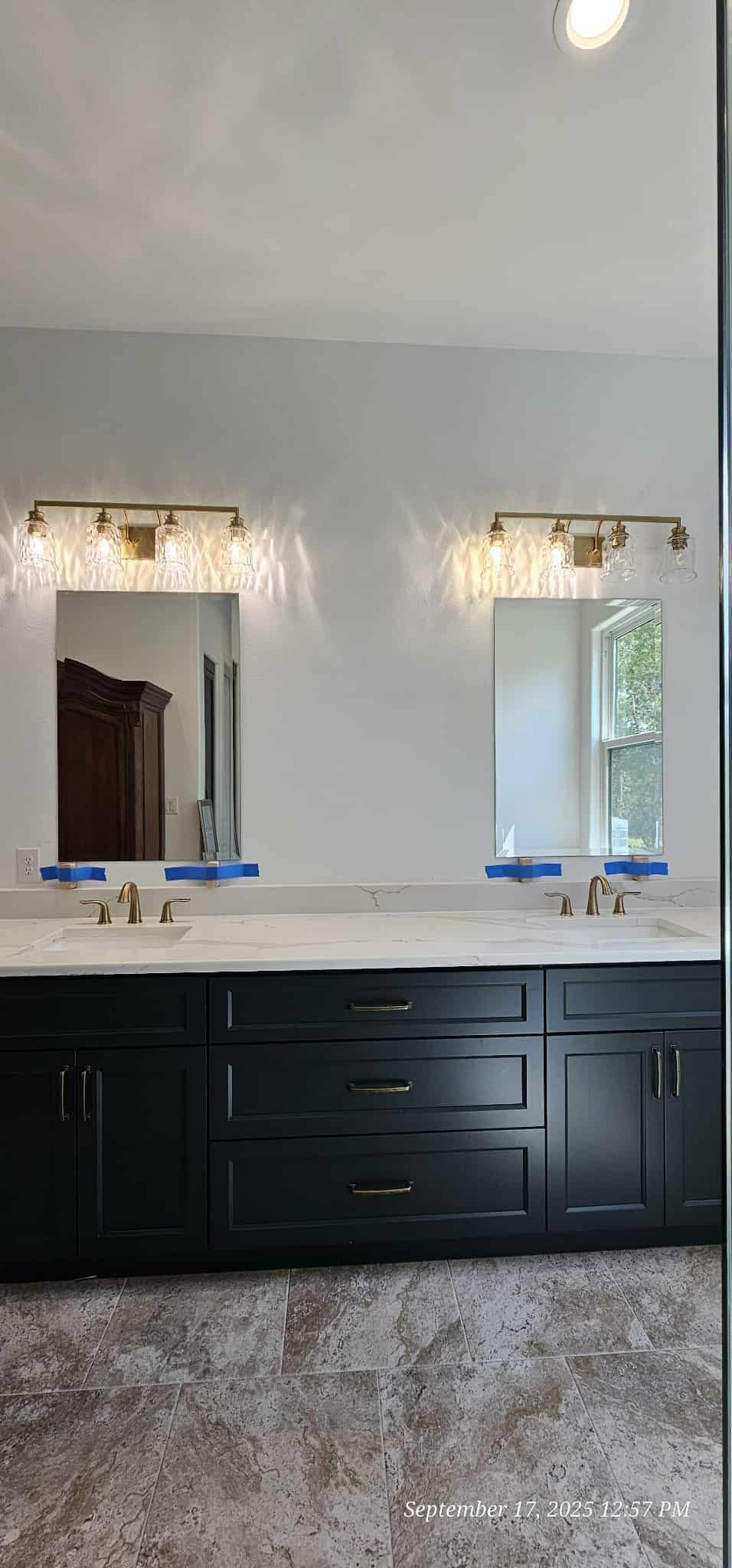 Bathroom with black cabinets, white countertop, two mirrors, and gold lighting fixtures.
