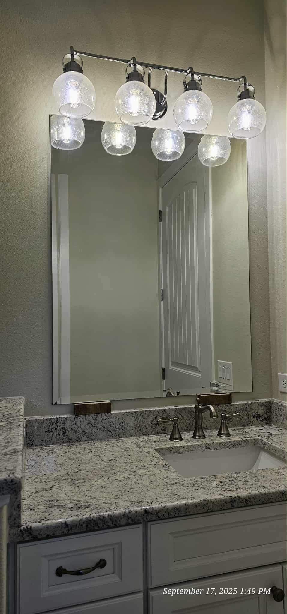 Bathroom vanity with mirror, overhead lights, and white cabinets. Gray granite countertop, sink, and faucet visible.