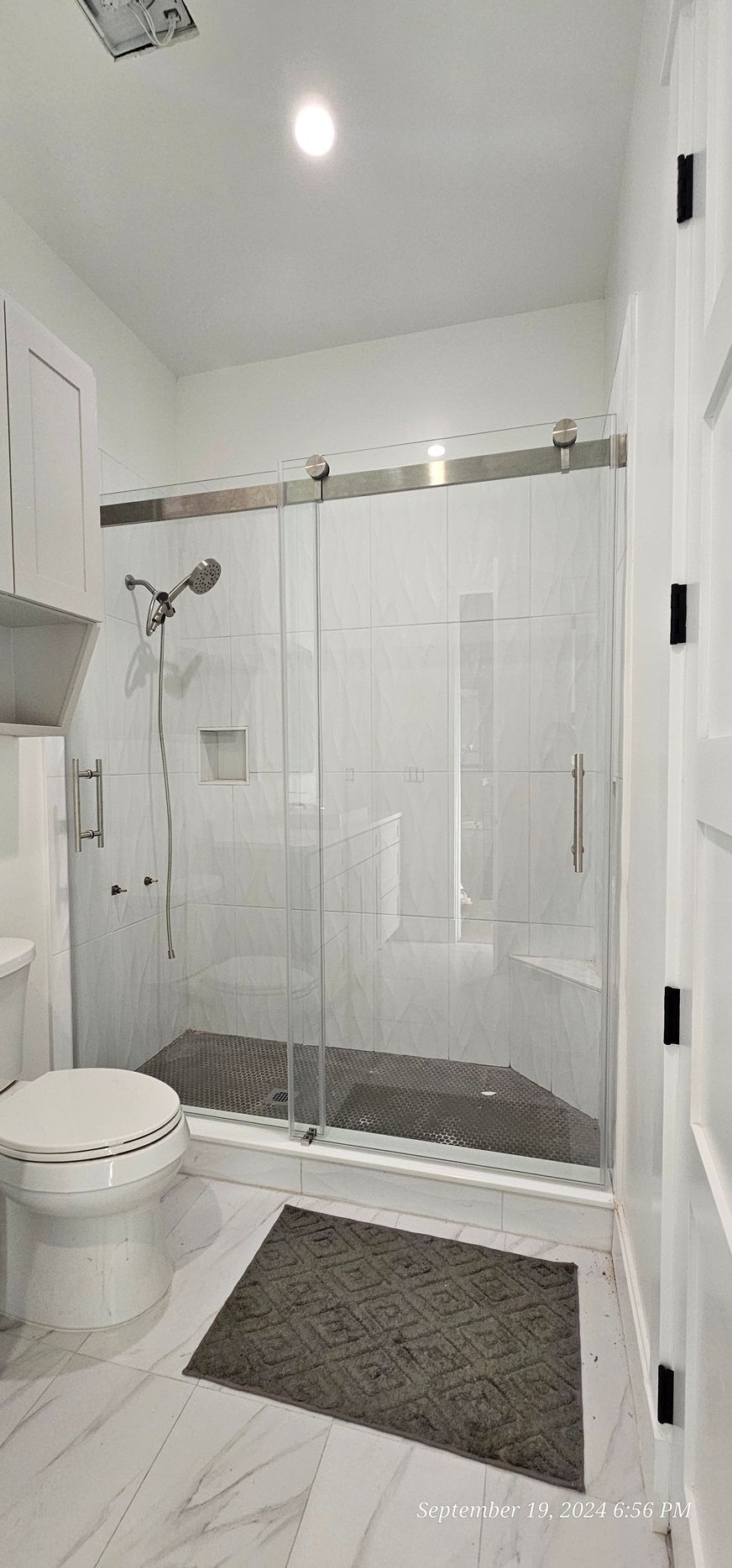 Bathroom with a glass shower, white toilet, and gray bath mat on a light-colored tile floor.