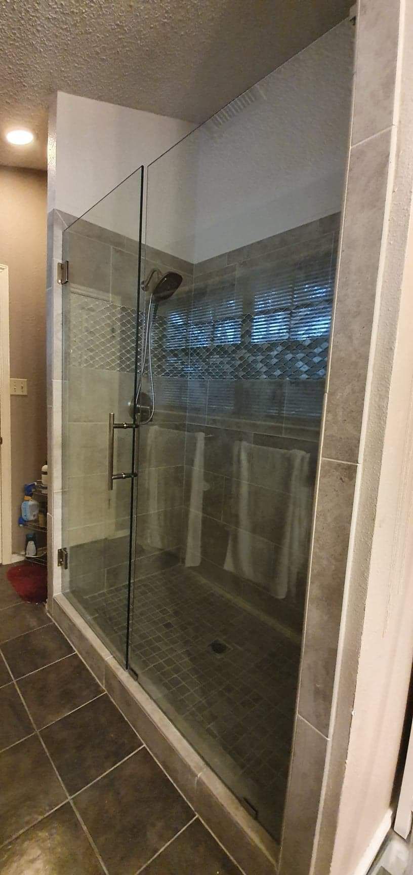 A glass shower enclosure with gray tile and a textured wall. Towels hang inside.