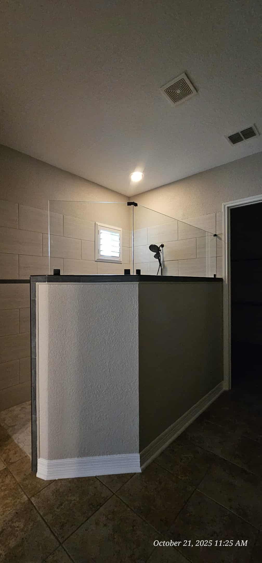 Shower stall with glass walls and light-colored tile.  Dark floor, black door, and a single ceiling light.