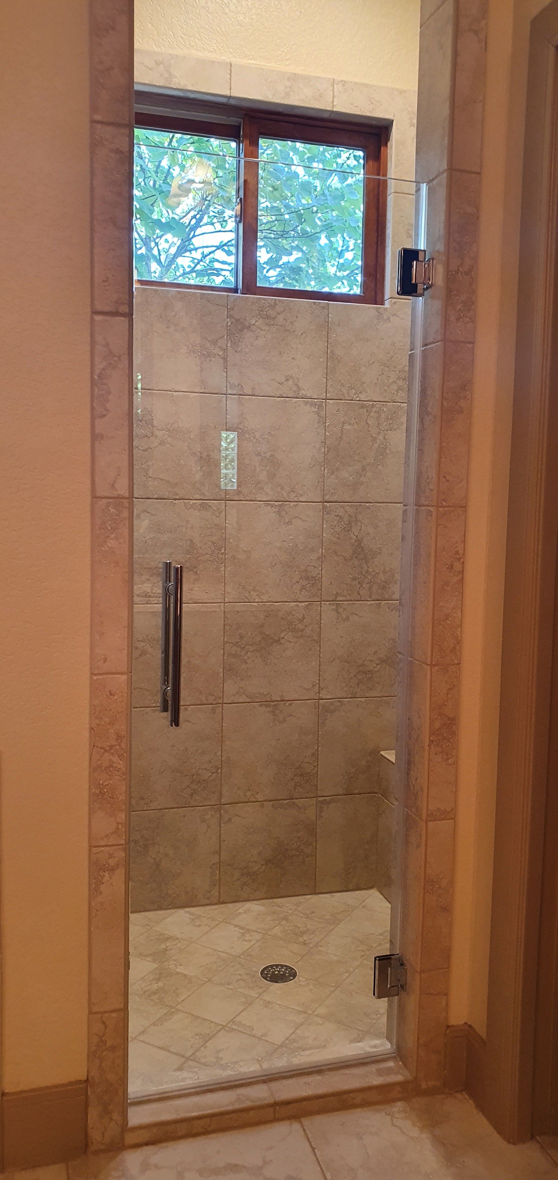 Shower stall with glass door and window above. Light tan tile with brown trim.