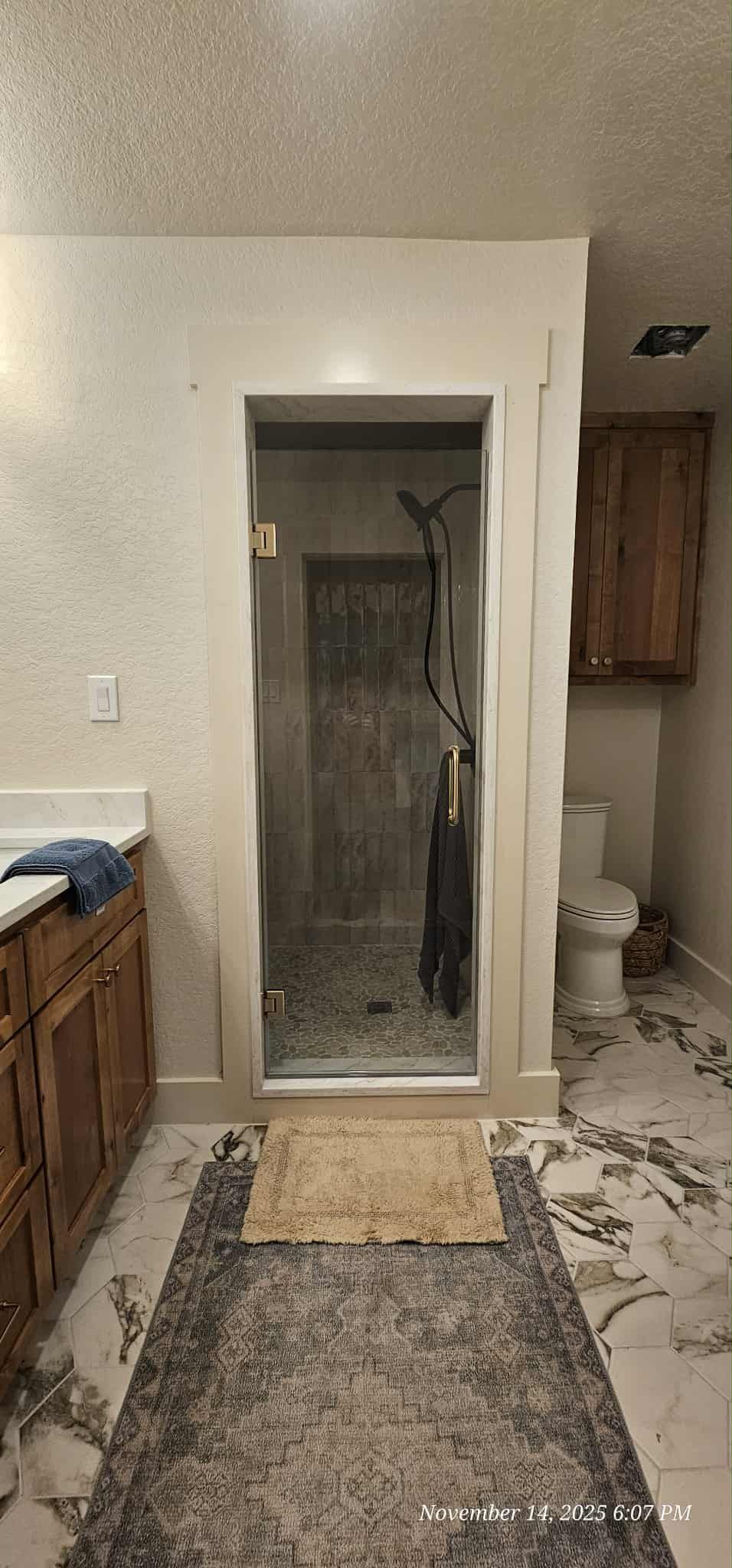 Bathroom with a glass shower, toilet, and wooden cabinets. The floor has a decorative rug.