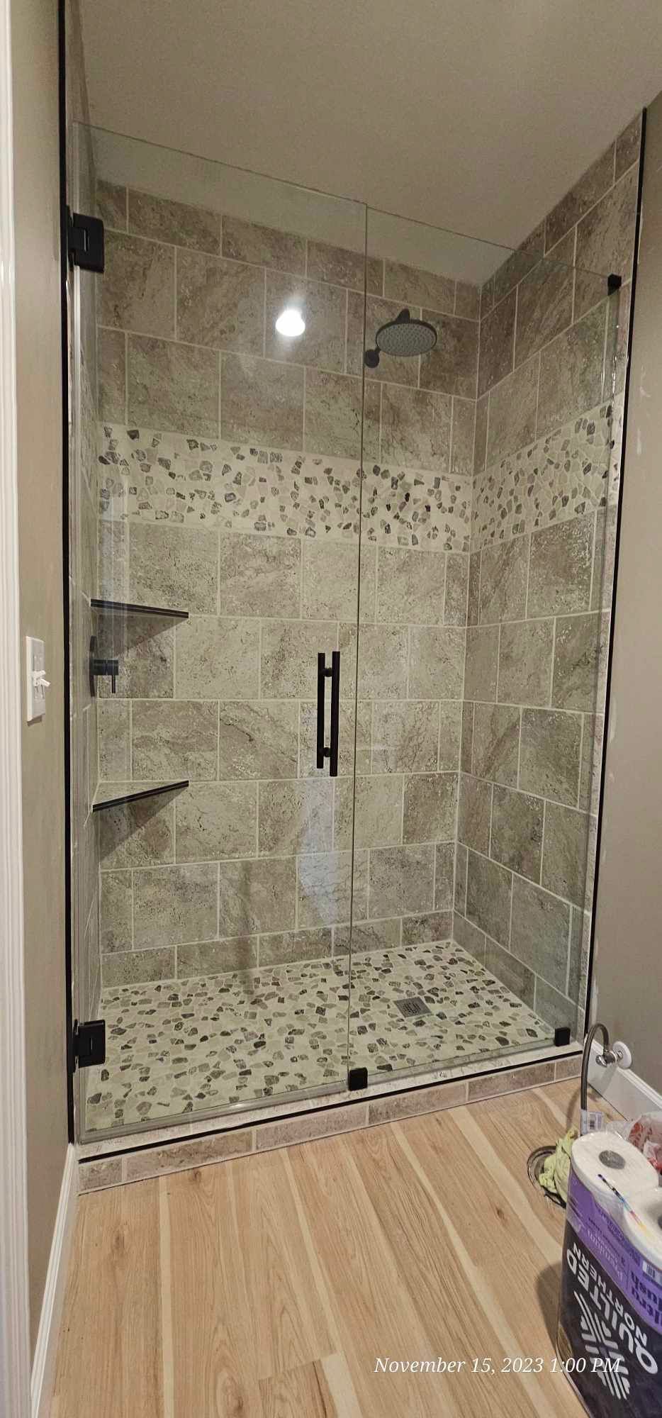A glass-enclosed shower with light-colored tile and wooden-look flooring. Two glass shelves are mounted inside.